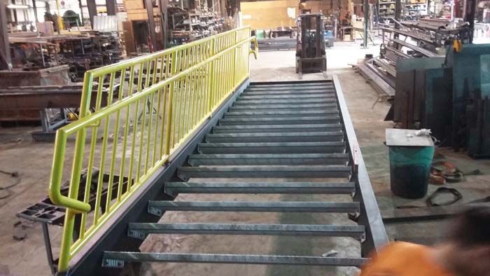 A flight of steel stairs with a bright yellow railing under construction in an industrial workshop.