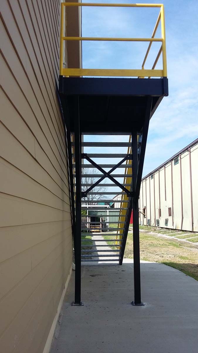 Exterior metal staircase with yellow railings attached to the side of a building, leading to a raised landing.
