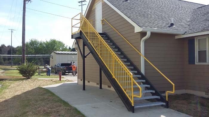 An exterior metal staircase with bright yellow railings leading to an upper-level doorway of a brown building.