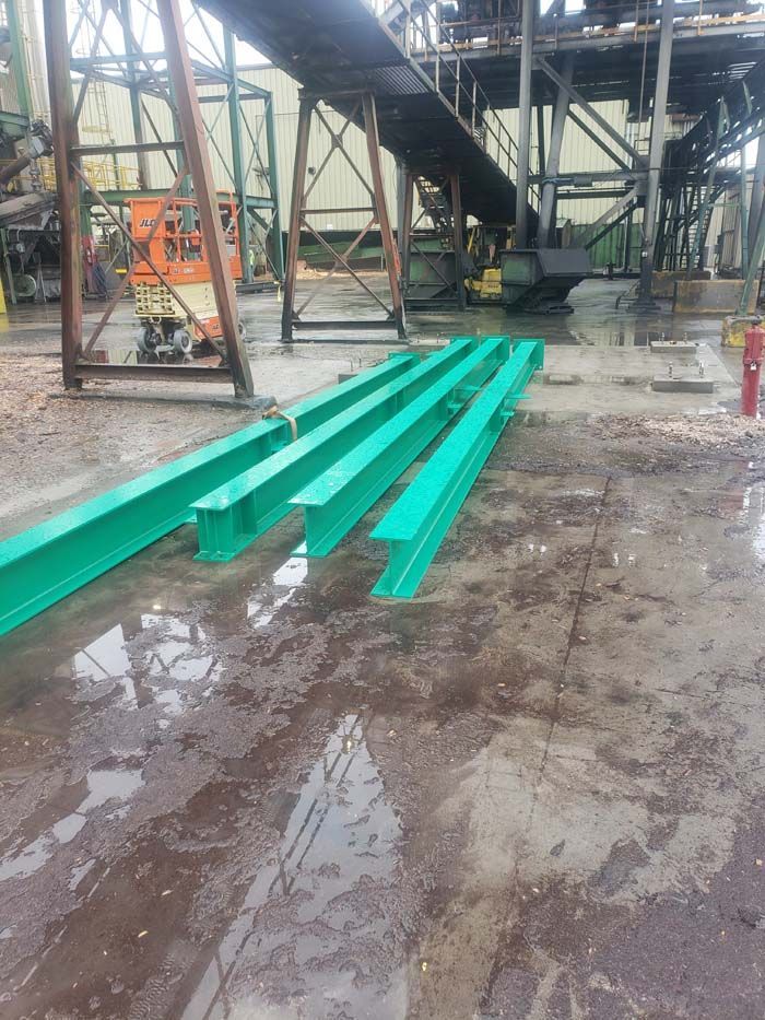 Three long green painted I-beams lying on a muddy ground at an industrial site with steel structures in the background.