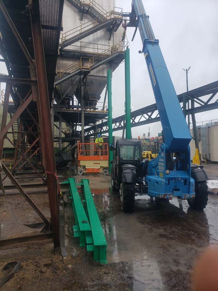 A blue telehandler lifts a green steel support beam at an industrial site with a scissor lift nearby.