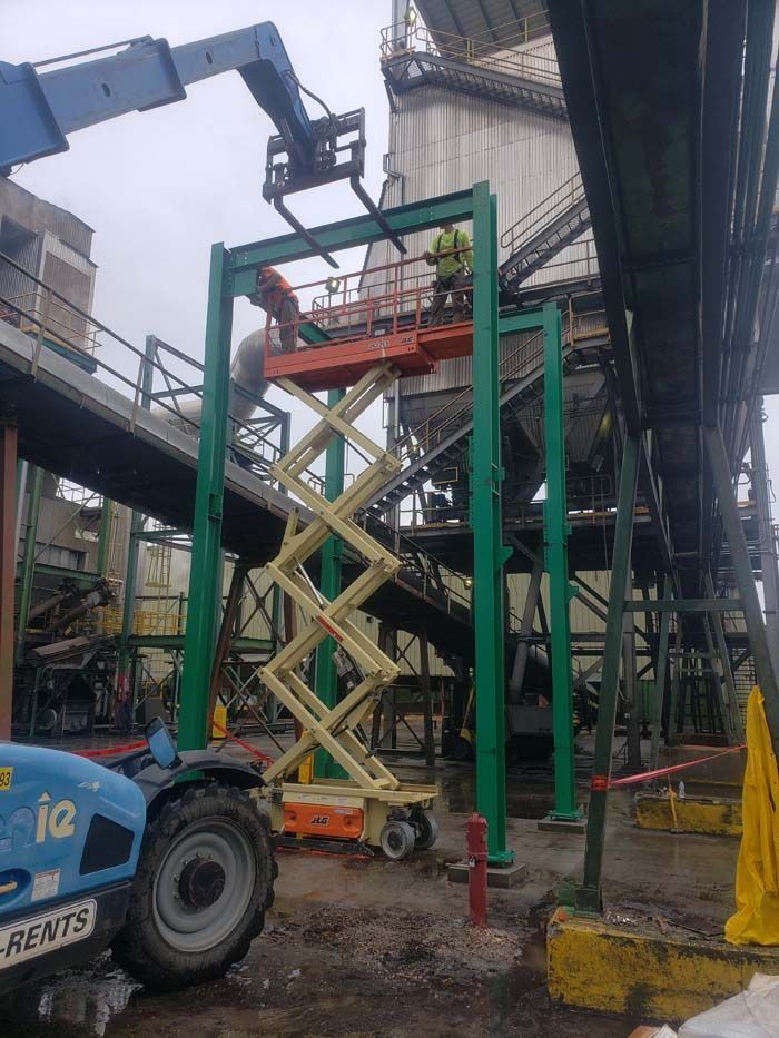 Workers stand on a scissor lift beneath a green metal frame at an industrial site as a telehandler crane arm hovers above.