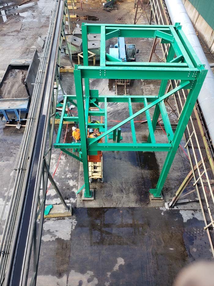A large, green, multi-level steel industrial frame stands on a concrete outdoor lot near a conveyor system.