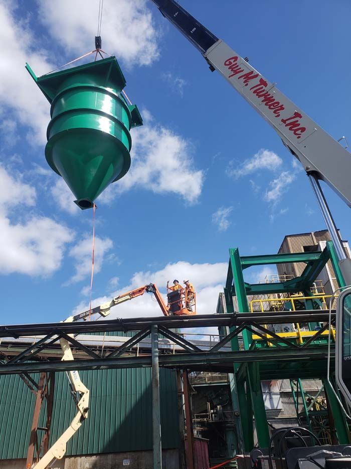 A crane lifts a large green conical hopper above a construction site with two workers in an aerial lift nearby.
