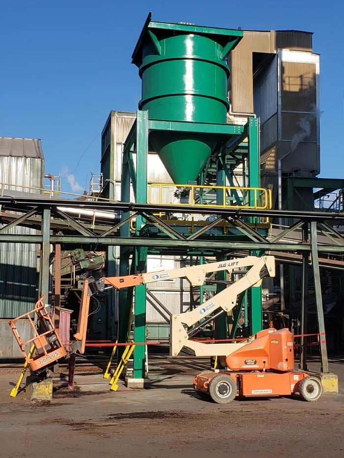 An orange boom lift stands in an industrial yard beneath a large, green metal hopper and overhead support structures.