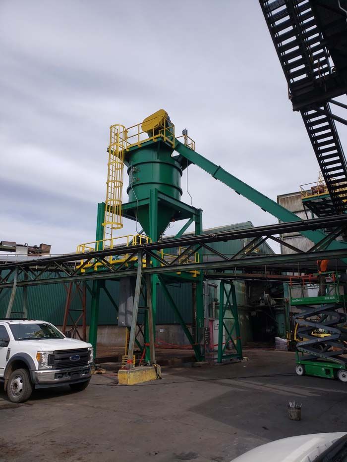 A tall green industrial silo with a yellow ladder and conveyor stands in a lot, beside a white pickup truck.