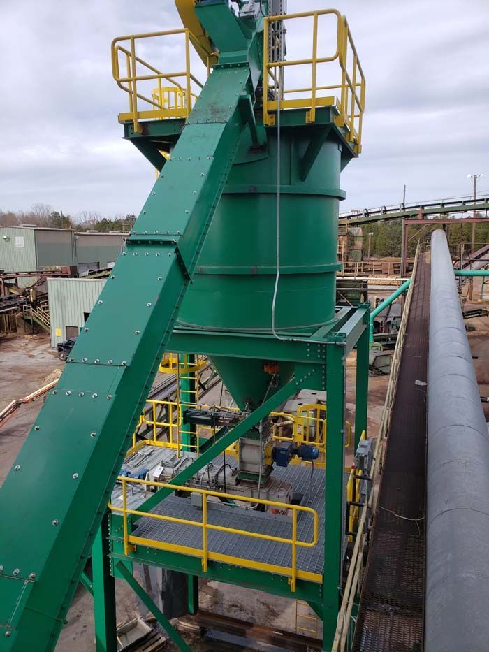 A large, industrial green storage hopper with a connected conveyor chute and yellow safety railings in an outdoor yard.