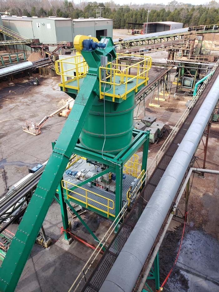 A green industrial storage silo with a conveyor system, elevated catwalks, and yellow railings in an outdoor plant yard.