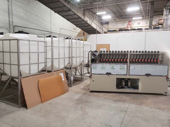 White industrial tanks in metal cages sit next to a machine with a row of circular components in a warehouse.