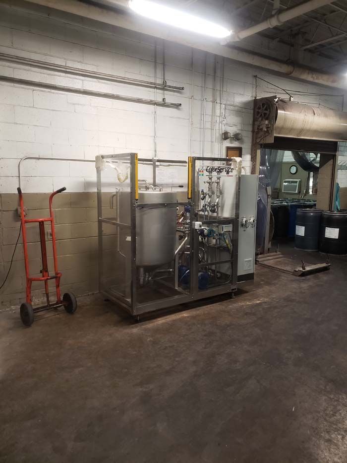 A metal industrial skid with a tank and plumbing inside a guarded frame, located in a warehouse near an orange hand truck.