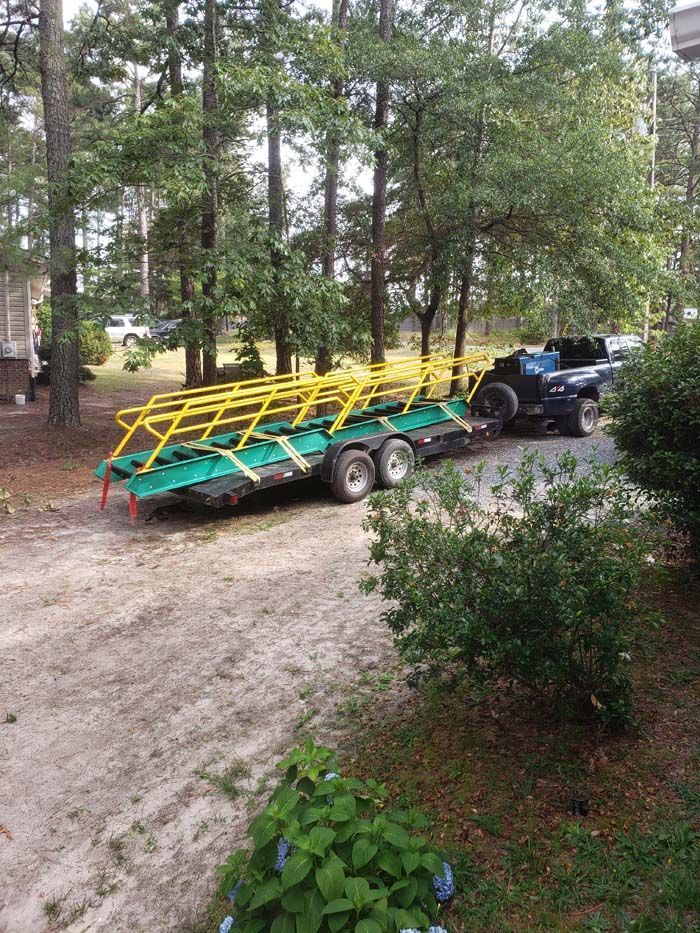 A pickup truck pulls a flatbed trailer loaded with green bench-style seating with yellow metal railings in a wooded yard.