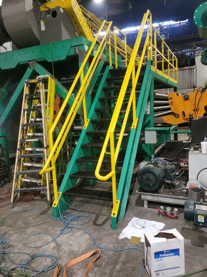 A metal industrial staircase with yellow railings and a green frame stands in a workshop next to a tall ladder.