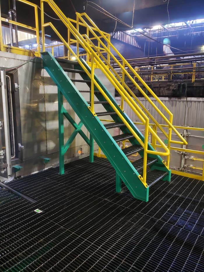 Industrial metal staircase with green stringers and bright yellow handrails on a grated platform in a factory setting.