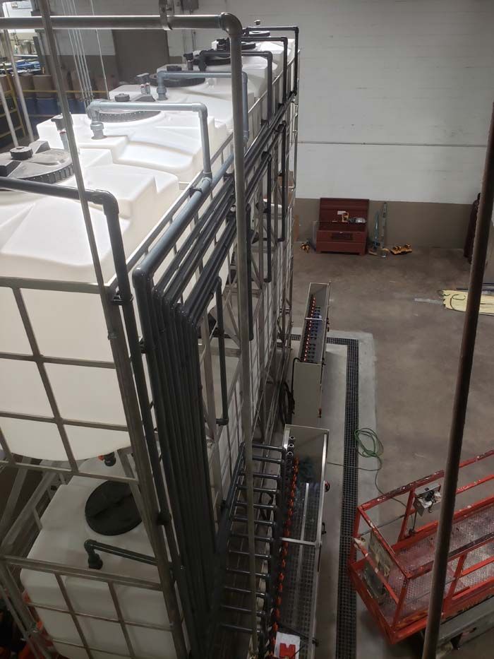 High-angle view of white industrial tanks in metal cages, connected by piping inside a warehouse with a red scissor lift.