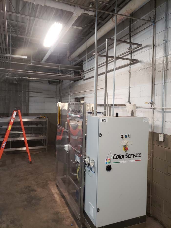 A ColorService industrial control unit stands in a workshop with exposed brick walls, plumbing pipes, and a stepladder.