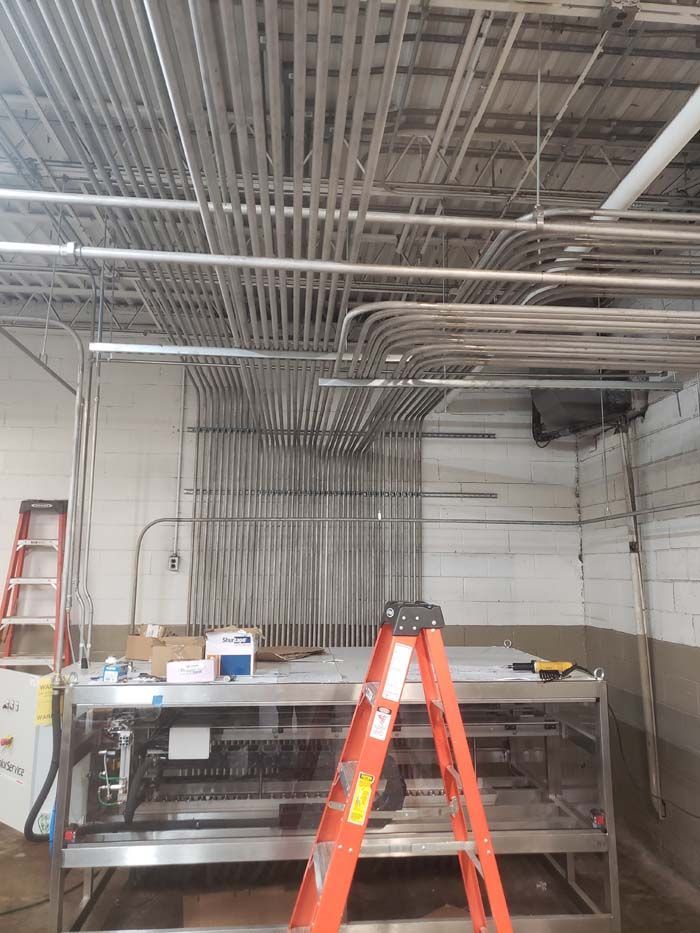 An orange stepladder sits before a large, stainless steel table in a utility room filled with numerous overhead conduits.