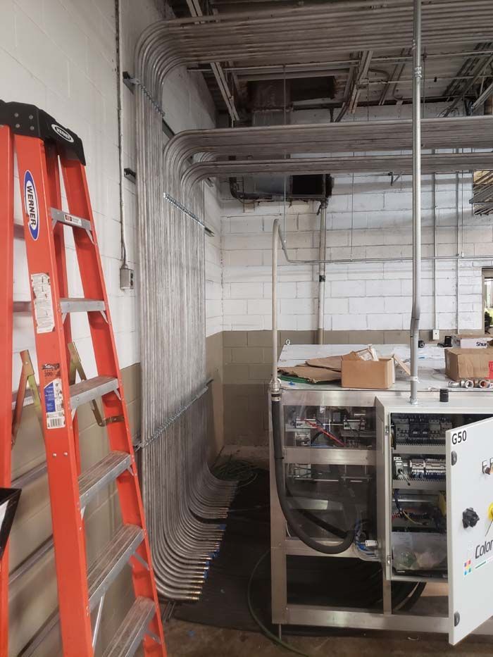 An orange ladder stands beside a wall with many metallic electrical conduits running into a piece of industrial equipment.