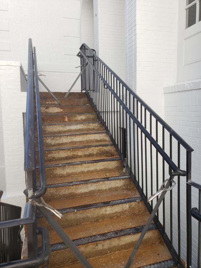 A set of outdoor concrete stairs with dark metal handrails, partially blocked by metal rods, against a white wall.