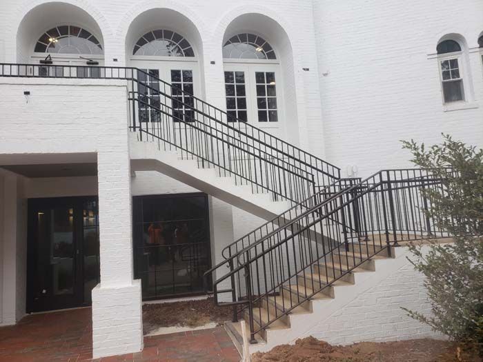 A white brick building with three arched doors on an elevated landing, accessed by a set of black metal stairs and railing.