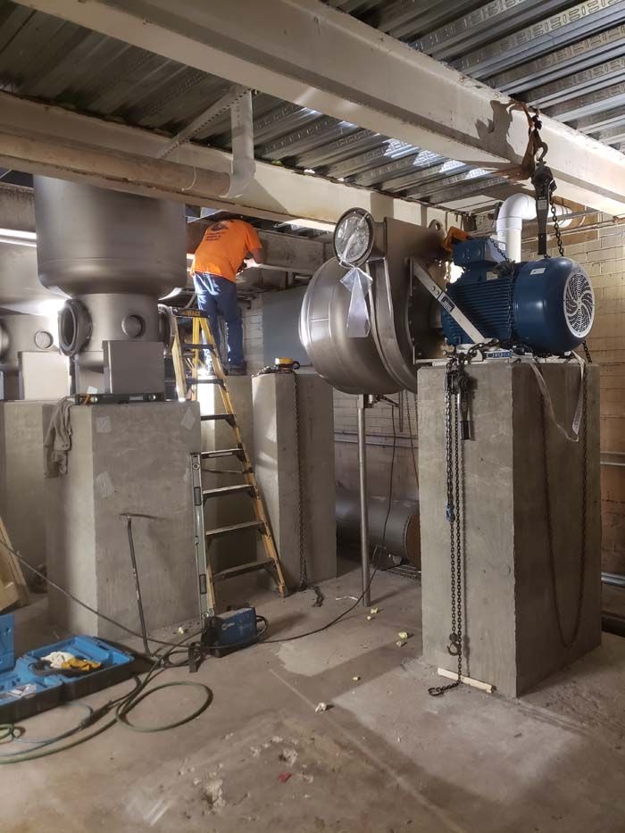 A worker in an orange vest on a ladder installing industrial machinery on concrete pedestals in a mechanical room.