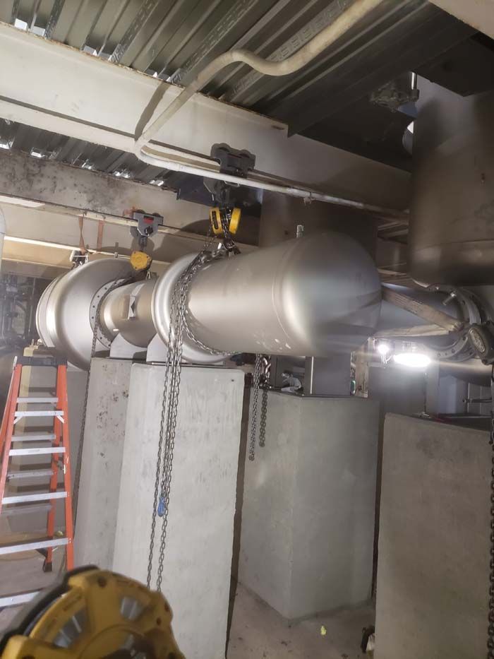A large metal cylindrical industrial tank suspended by chains and hoists above concrete supports in a mechanical room.