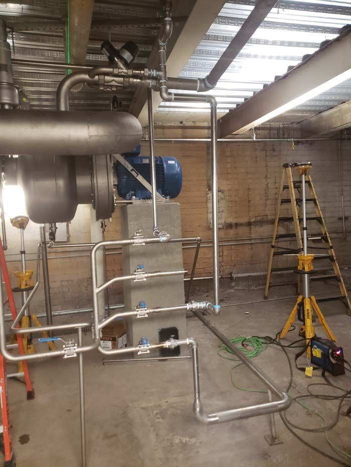 Stainless steel piping system under construction in an industrial facility with a step ladder and equipment in the room.