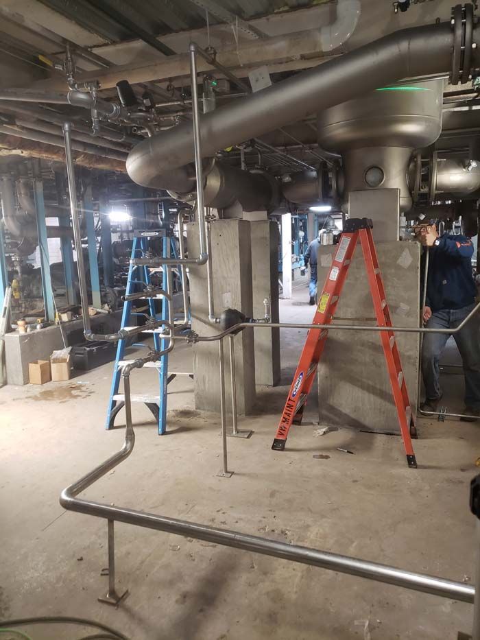 A person working near complex stainless steel piping in an industrial construction site with two ladders in the area.