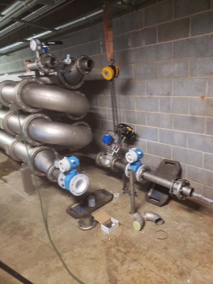 Industrial plumbing setup with large metal pipes, blue control valves, and a chain hoist against a grey brick wall.