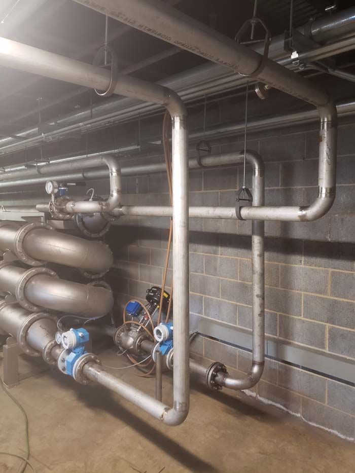 Industrial pipework featuring metal tubes, valves, and gauges mounted against a grey cinder block wall.