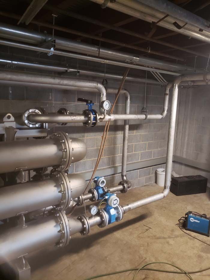 Industrial piping system with stainless steel pipes, valves, and blue pressure sensors against a concrete block wall.