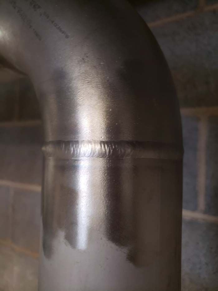 A close-up of a neat, uniform metal weld connecting a pipe elbow to a straight section of stainless steel pipe.