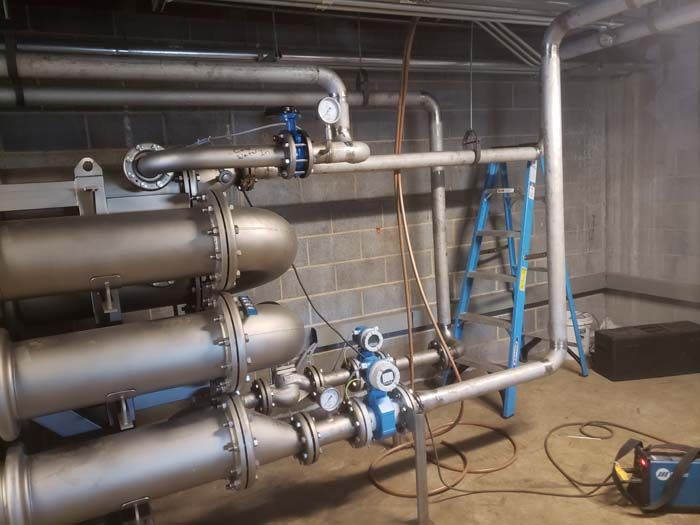Industrial pipes with valves and gauges in a concrete-walled mechanical room, with a ladder and welding equipment nearby.