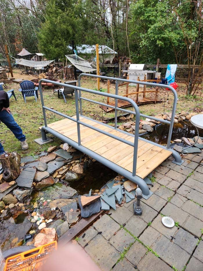 A newly installed, gray metal-framed garden bridge with wooden planks crossing over a small, stone-lined backyard pond.