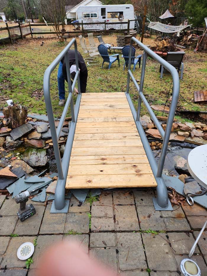 A newly installed wooden footbridge with grey metal railings sits on a stone patio spanning a garden pond.