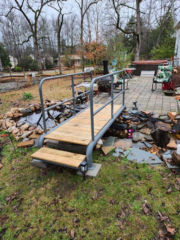 A wooden garden bridge with gray metal railings spanning a small rocky pond, leading to a paved patio area.
