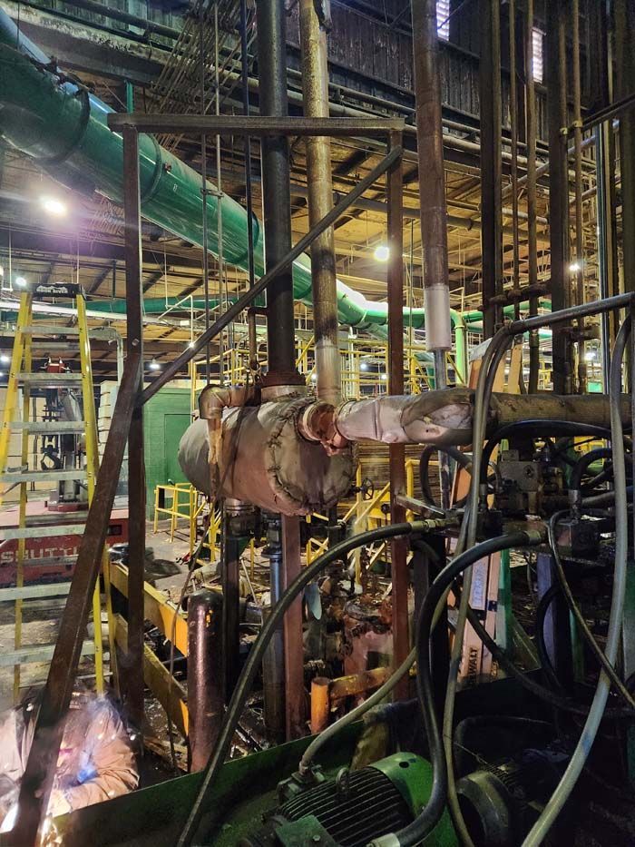 A worker welds a pipe in an industrial facility surrounded by metal structures, machinery, and a yellow stepladder.