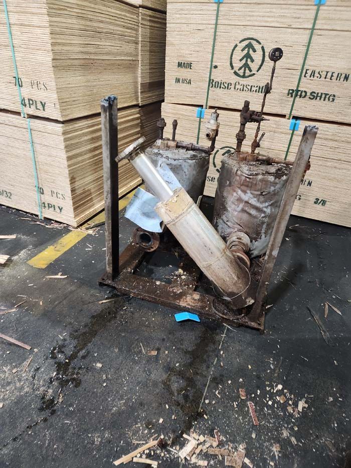 An old, weathered industrial pump unit with metal cylinders and pipes sitting on a concrete floor near wood crates.
