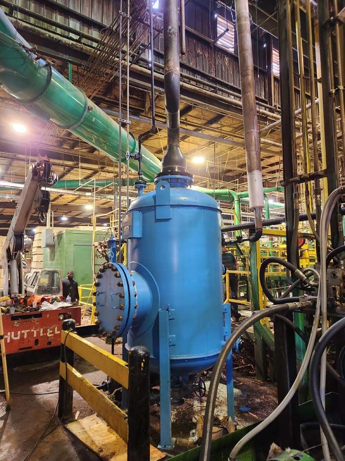 A large blue industrial tank stands upright in a factory, connected to overhead pipes with a flanged side access port.