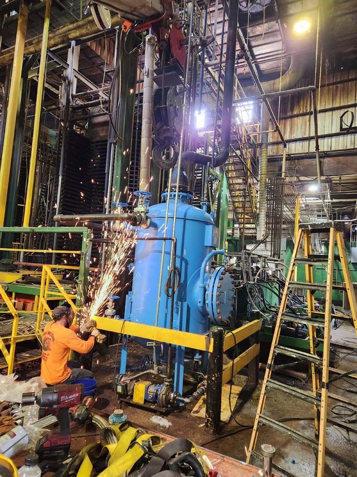 A worker in an orange shirt uses an angle grinder on a metal pipe attached to a large blue tank in an industrial space.