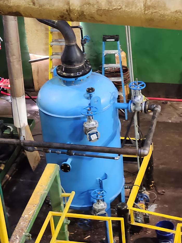 A large, blue industrial pressure vessel stands on a factory floor, connected to various pipes, valves, and a ladder.
