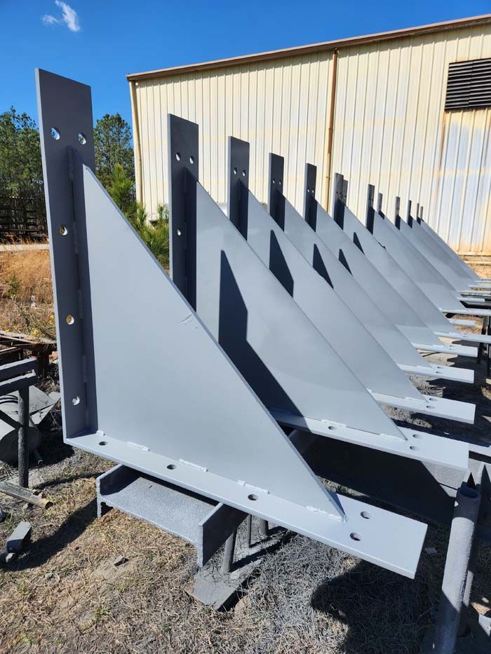 A row of gray, triangular steel support brackets lined up outdoors on a gravel lot under a clear blue sky.