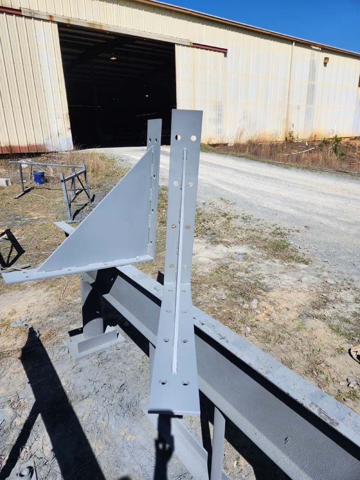 Two grey metal L-shaped brackets with welded gussets resting on a steel beam outside a large metal industrial building.
