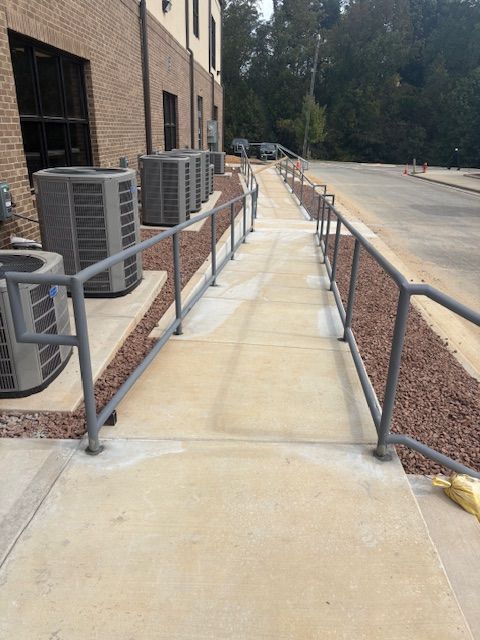 An outdoor concrete wheelchair ramp with gray metal handrails running alongside a brick building and HVAC units.