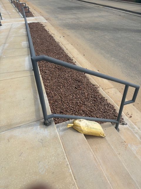 A gray metal handrail runs along stone stairs next to a gravel garden, with a yellow sandbag lying on the bottom step.