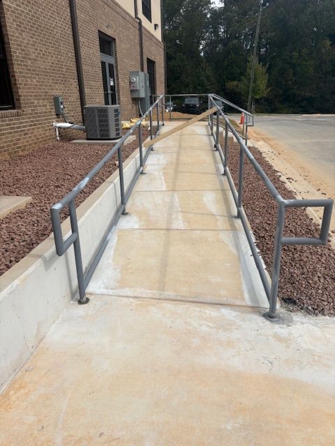 Concrete wheelchair ramp with grey metal railings, partially blocked by a wooden board, leading to a brick building.