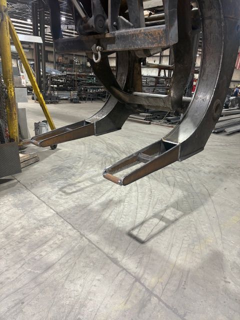 A close-up of a heavy-duty industrial metal grapple attachment suspended in a workshop setting.