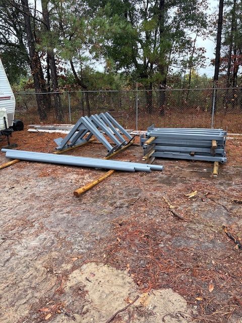 Steel framing components for a building structure, including triangular trusses and long beams, on a dirt ground outdoors.