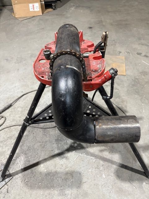 A red chain vise mounted on a tripod stand, holding a black metal elbow pipe connected to a straight pipe section.