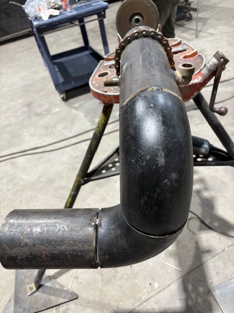 A black steel pipe elbow is clamped into a metal pipe stand in a workshop, positioned for welding or assembly.