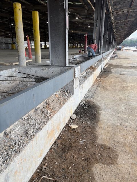 A person works on repairs at a construction site with exposed concrete footings and steel support columns.